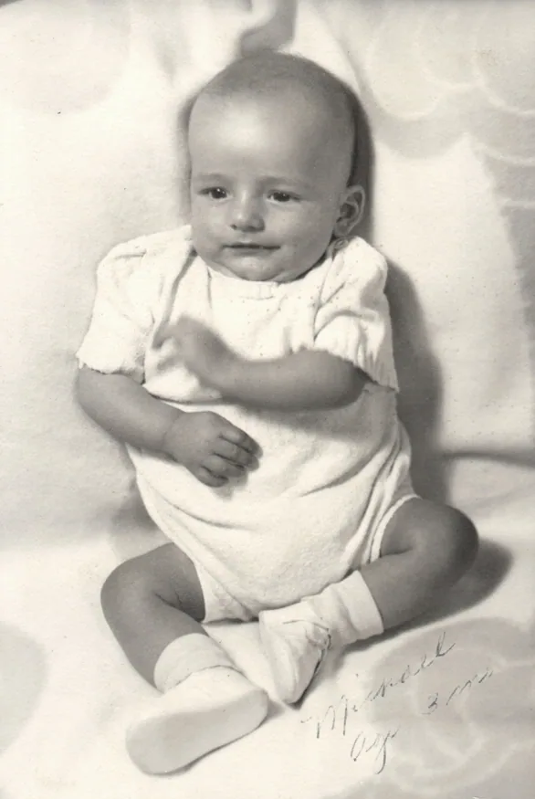 Black and white photograph of a smiling infant sitting on white fabric wearing a light-colored dress with short sleeves and white socks