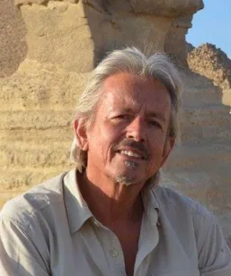 Older man with white hair and mustache smiling at camera, wearing light colored shirt outdoors with stone structure in background
