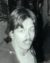 Black and white portrait of a man with shoulder-length hair and a mustache wearing a dark shirt