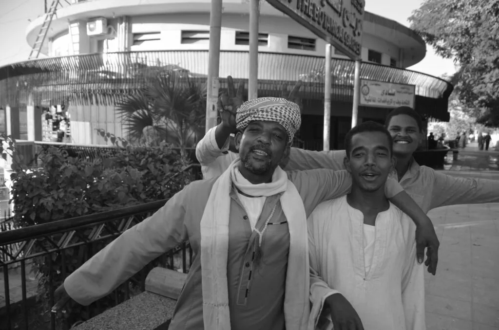 Three men posing together on a street in front of a modern circular building, black and white photograph