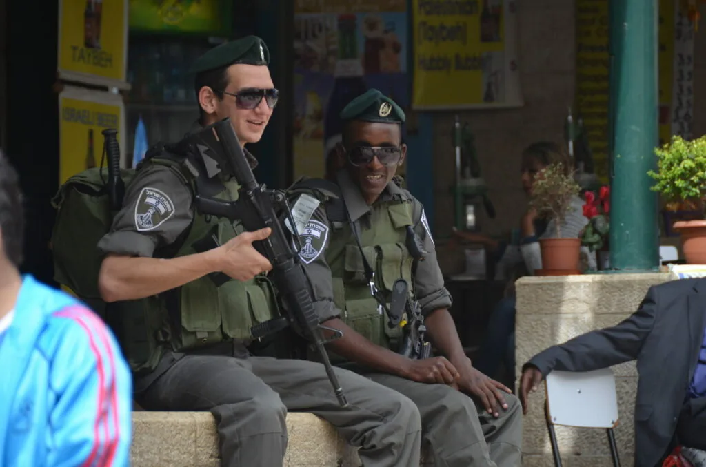 Two armed security personnel in tactical gear sitting on a bench with yellow signage visible in the background