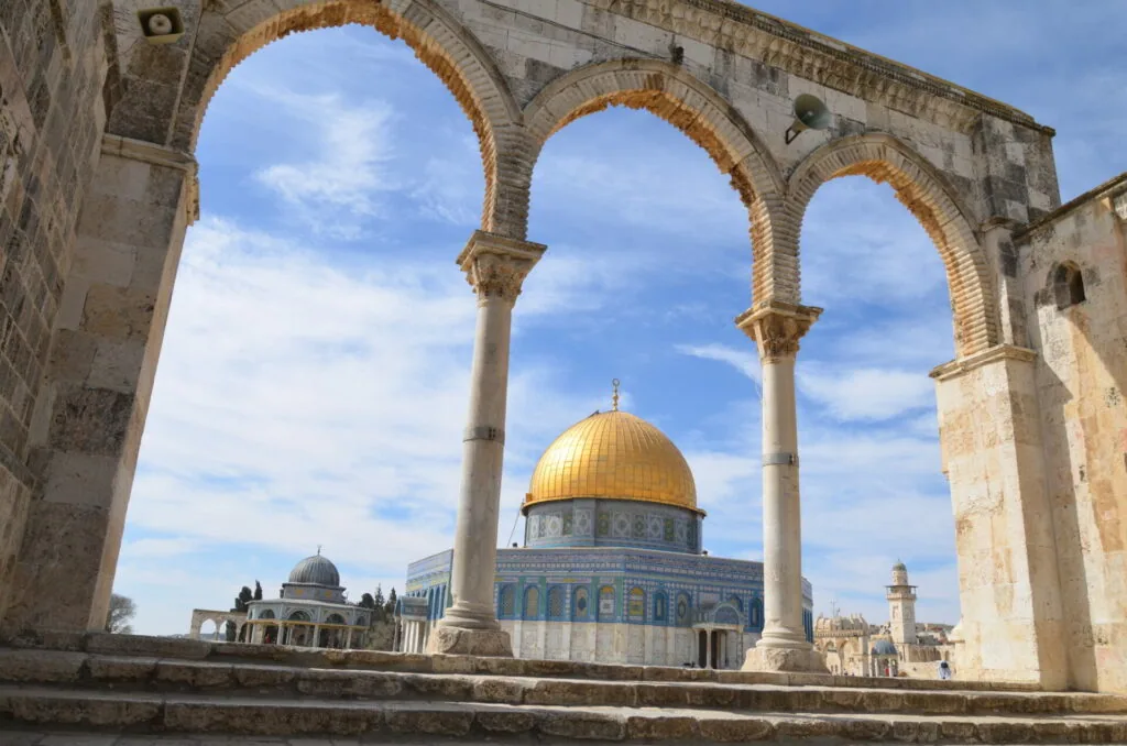 Golden dome building framed through three stone arches with blue sky and clouds visible