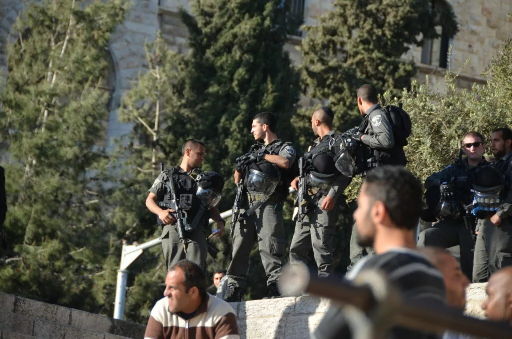 Armed law enforcement officers standing in formation near cypress trees and stone structures, with civilians in foreground.