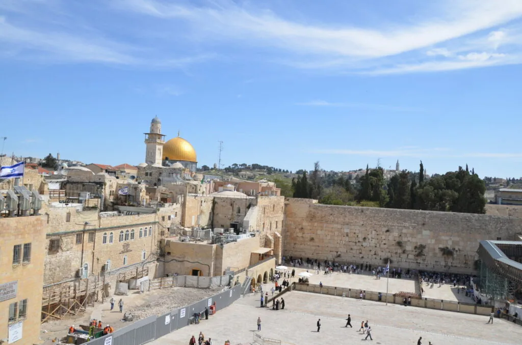 Western Wall plaza with golden Dome of the Rock mosque in the distance, stone walls, cypress trees, and visitors