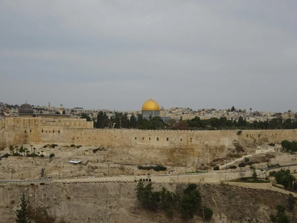 Ancient stone walls surrounding a hillside city with a golden domed religious structure and dense urban buildings in the background under overcast sky