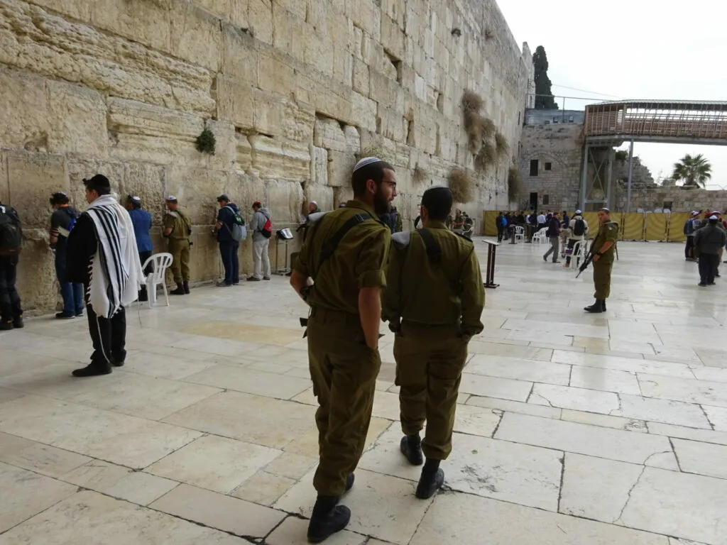 Two uniformed soldiers walking in front of an ancient stone wall with visitors gathered nearby