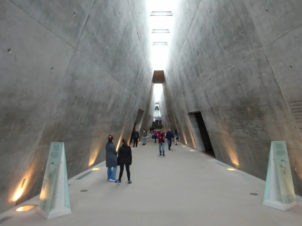 Long concrete corridor with high walls, skylights overhead, and visitors walking toward distant exit