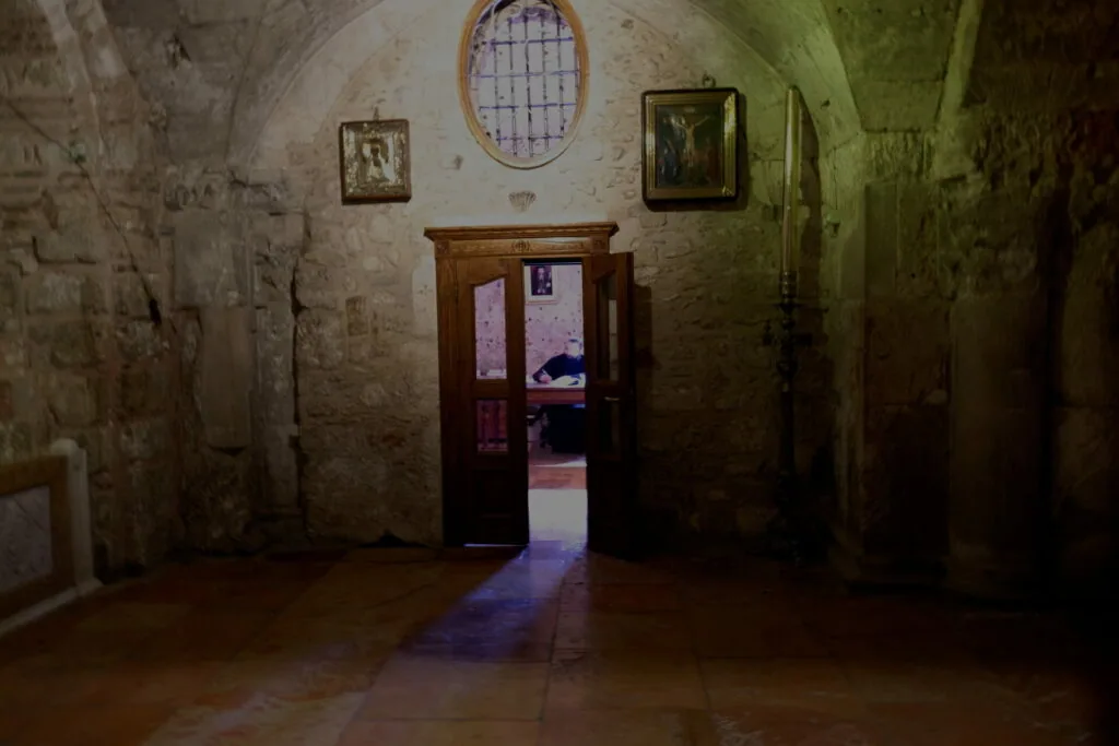 Dimly lit stone interior room with wooden door, oval window, framed pictures on walls, and colored light casting shadows on floor