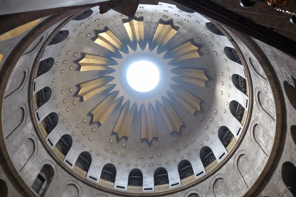 Circular architectural dome with radiating ribbed sections, arched windows around the base, and bright light streaming through the center opening.