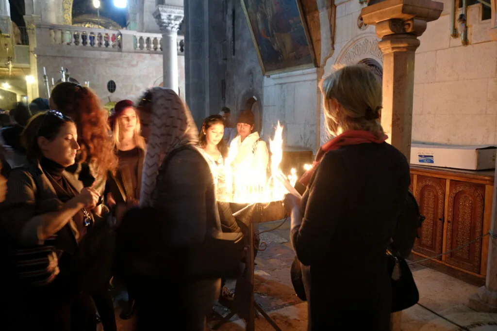 Group of people gathered around a large flame-filled vessel in a stone courtyard at night near a wooden door and ornate pedestal.