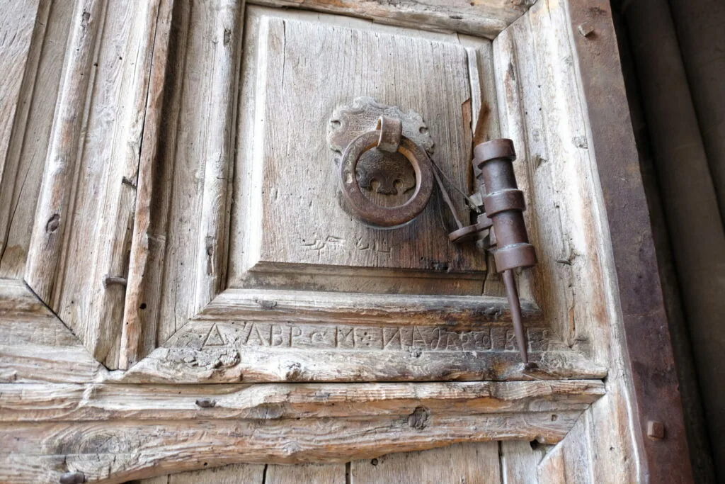Weathered wooden door panel with ornate iron ring knocker and latch, showing carved text inscription