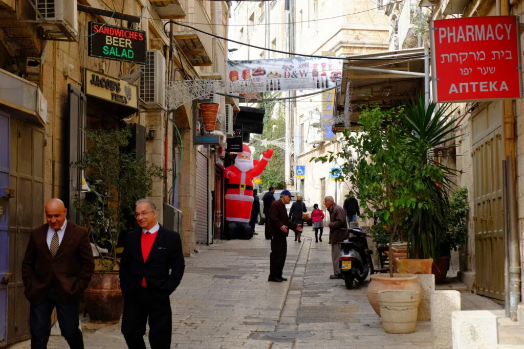 Narrow alleyway in Jerusalem's Old City with pedestrians, stone buildings, shops, and a large inflatable Santa decoration