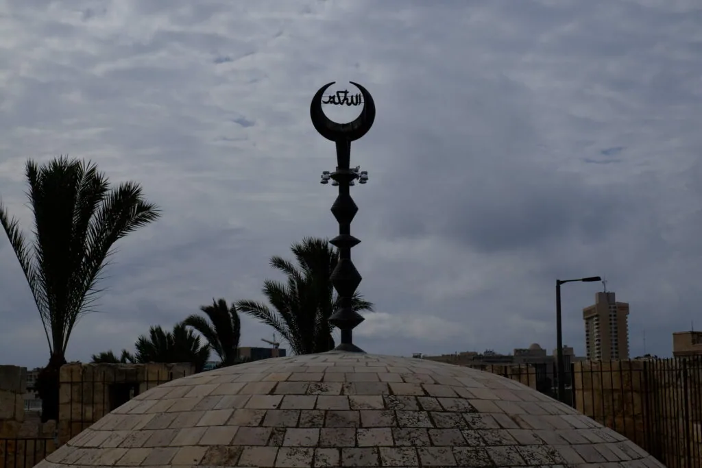 Ornamental Islamic finial with crescent and cross symbol atop domed structure with palm trees and urban buildings in background