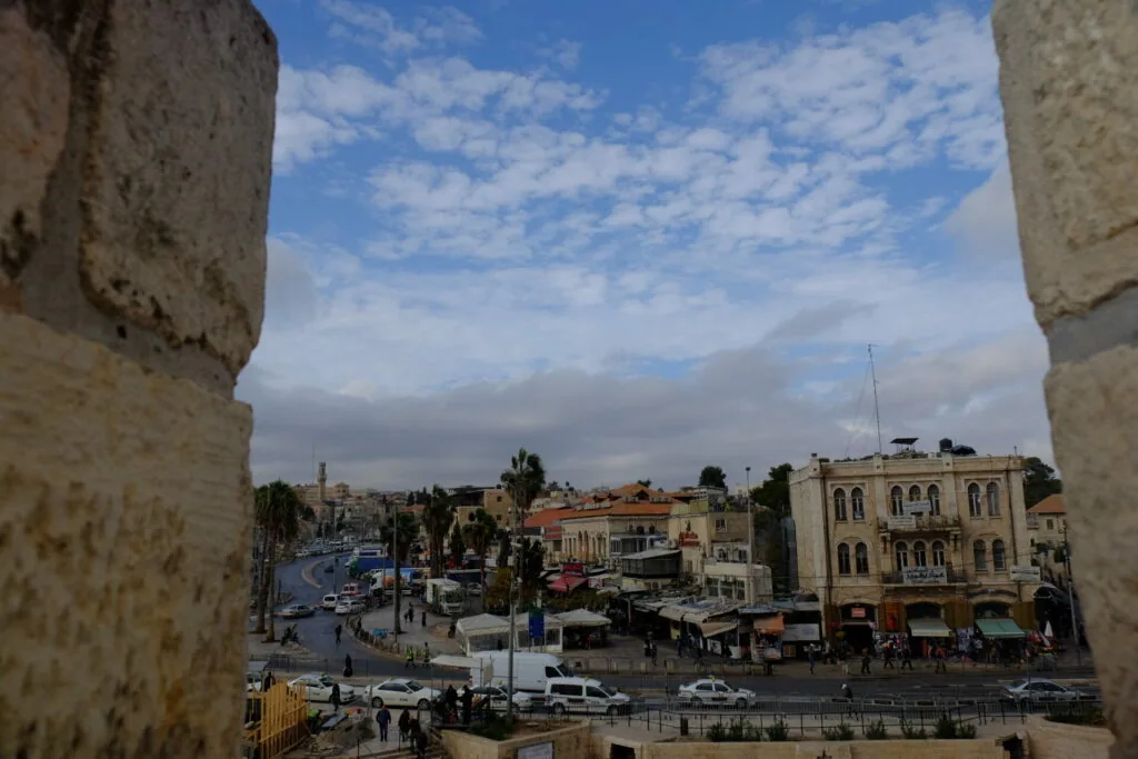 View through stone archway of Middle Eastern city square with historic buildings, palm trees, and blue sky with white clouds