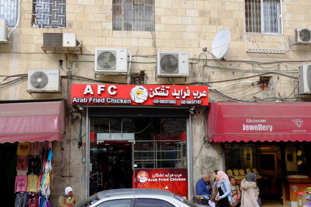 Arab Fried Chicken storefront with red signage and Arabic text on street with adjacent jewelry shop and residential building above