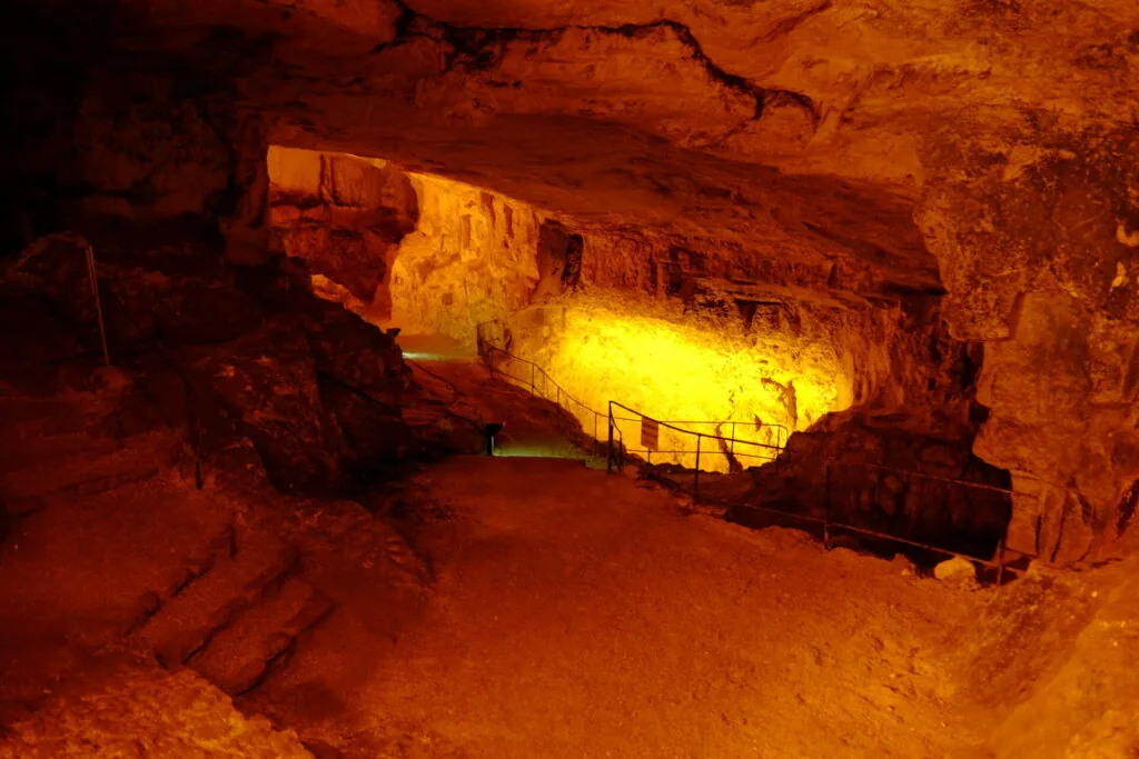 Underground cave chamber with golden yellow artificial lighting illuminating rock formations and a metal railing walkway