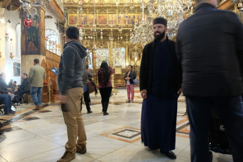 Interior of ornate Orthodox church with gold iconostasis, chandelier, and visitors walking on marble floor