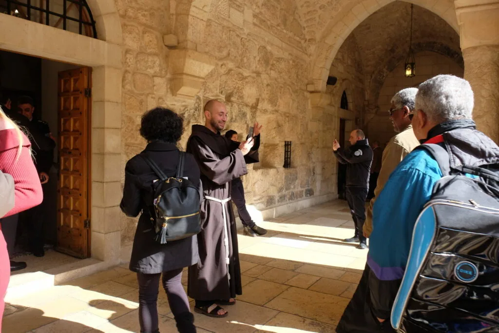 A robed guide gestures while speaking to tourists in a stone-vaulted corridor with arched passages inside a historic building.