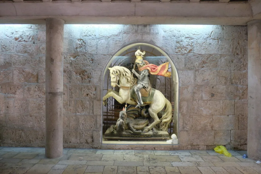 Relief sculpture of armored knight on white horse slaying dragon, mounted in arched stone niche within vaulted stone corridor