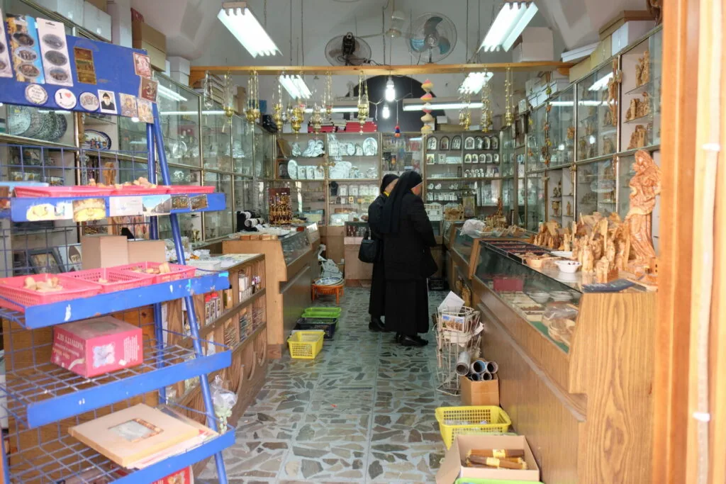 Small shop interior with glass display cases containing ceramics and decorative items, shelves with merchandise, and a woman in black clothing browsing.