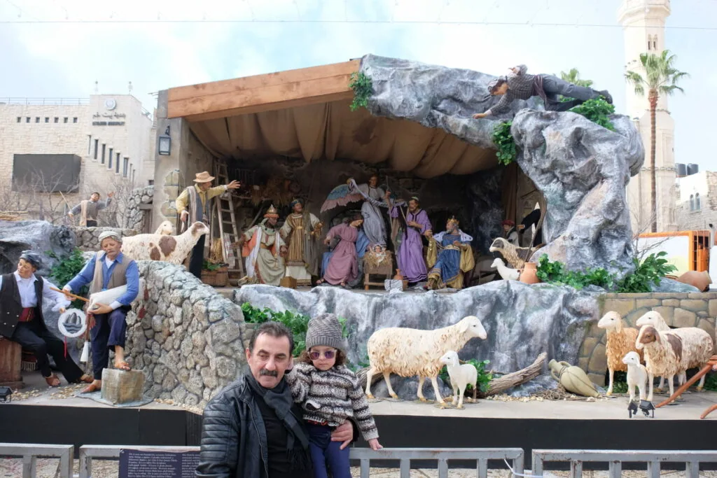 Man and child standing in front of large nativity scene display with figures, animals, and stone cave structure in urban setting