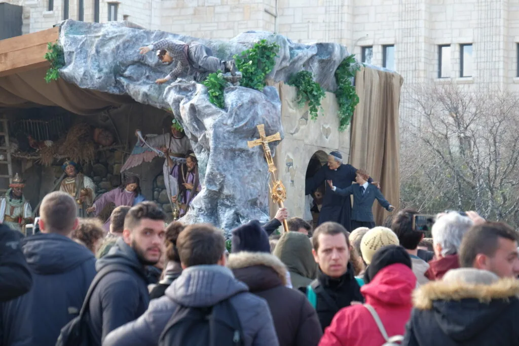 Crowd gathered at outdoor religious ceremony with large stone cross monument and nativity scene shelter