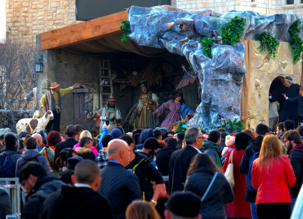 Crowd gathered around outdoor nativity scene stage with figures and painted backdrop of cave structure