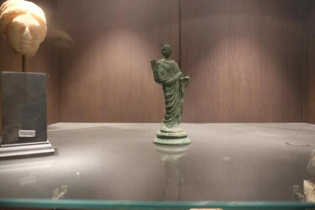 Green bronze statue of robed figure on pedestal in museum display with bronze head sculpture and textured wall backdrop