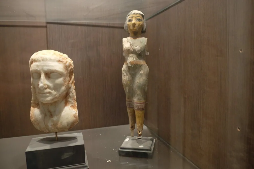 Ancient Egyptian stone sculptural head and figurine displayed in a museum case with brown wooden walls