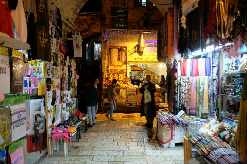 Narrow alleyway in a Middle Eastern market with illuminated storefronts, colorful textiles, and merchandise displayed on walls and ground.