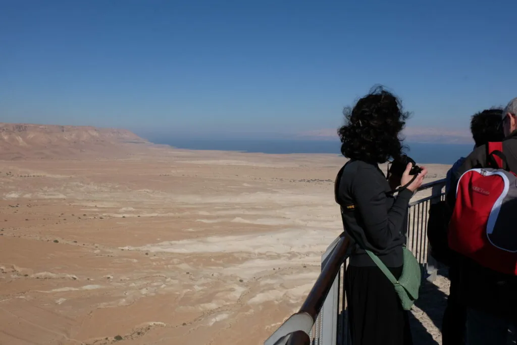 Tourists at elevated viewpoint overlooking arid desert landscape with body of water visible in distance under clear blue sky