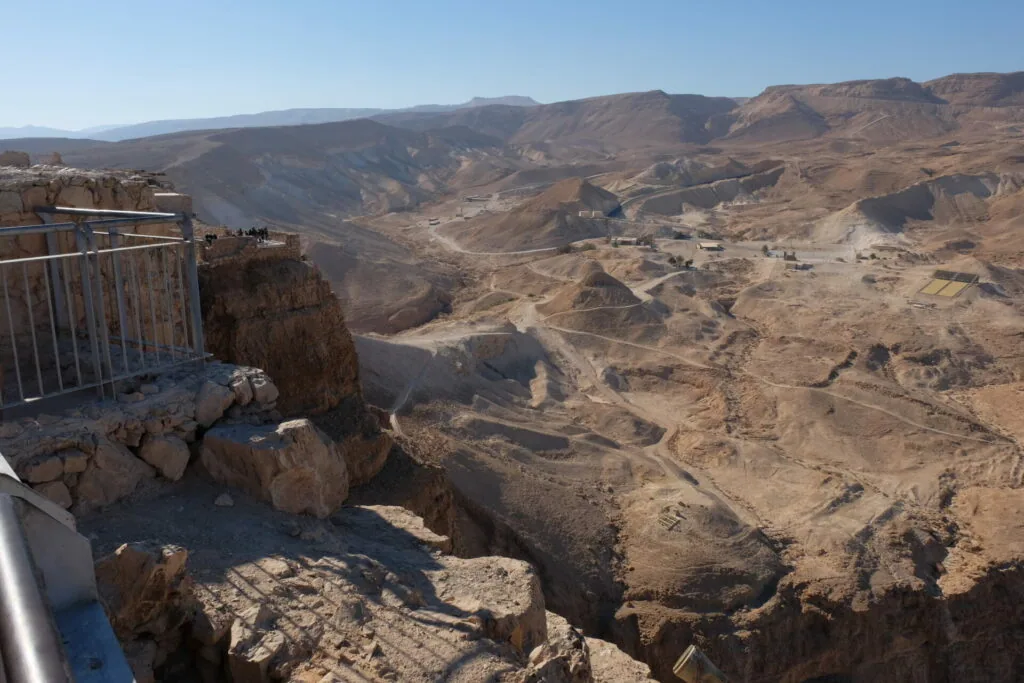 Arid desert landscape with rocky terrain, winding roads, and scattered settlements viewed from a high cliff overlook with metal railing in foreground
