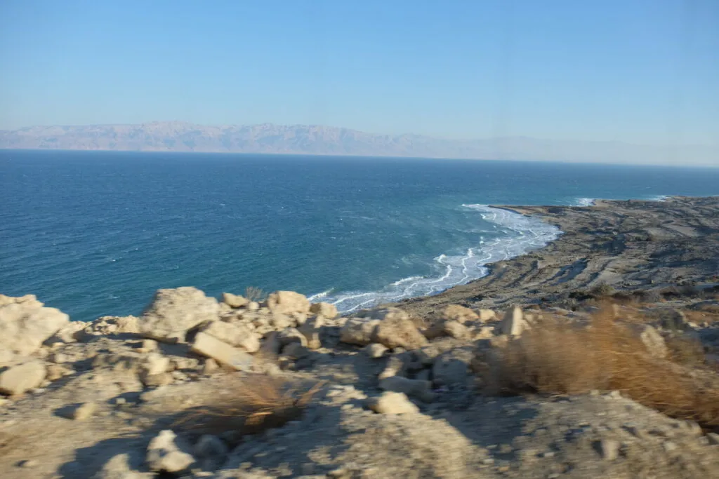 Rocky coastal cliffs overlook turquoise waters with waves breaking on shore and mountains visible across the sea horizon