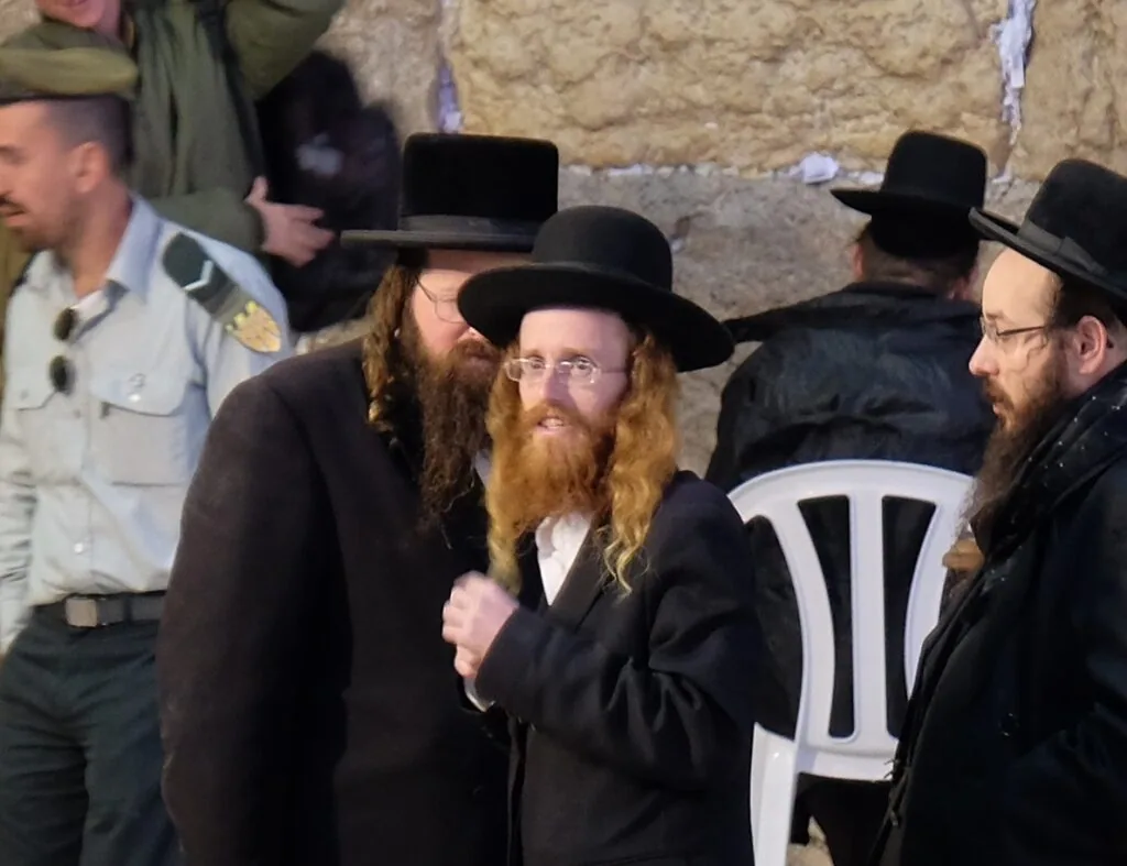Men with long beards wearing black hats and coats gathered at a stone wall during a religious ceremony