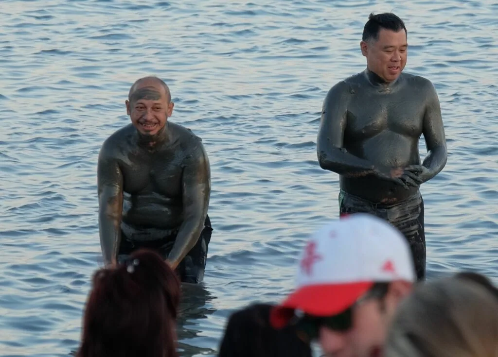 Two men covered in dark mud standing in shallow water, with spectators visible in the foreground