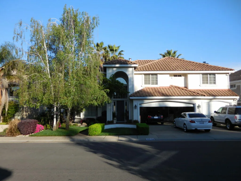 Two-story white stucco house with brown tile roof, two-car garage, and manicured front yard with weeping willow and pink flowering shrubs