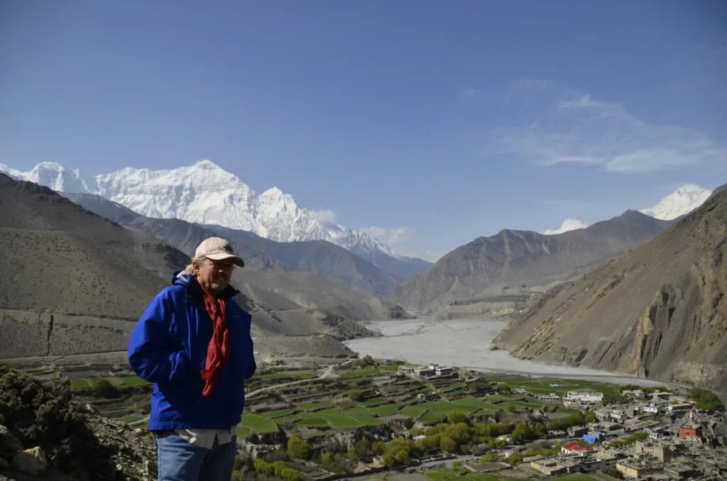 Person in blue jacket standing on mountainside overlooking valley village with snow-capped peaks in background