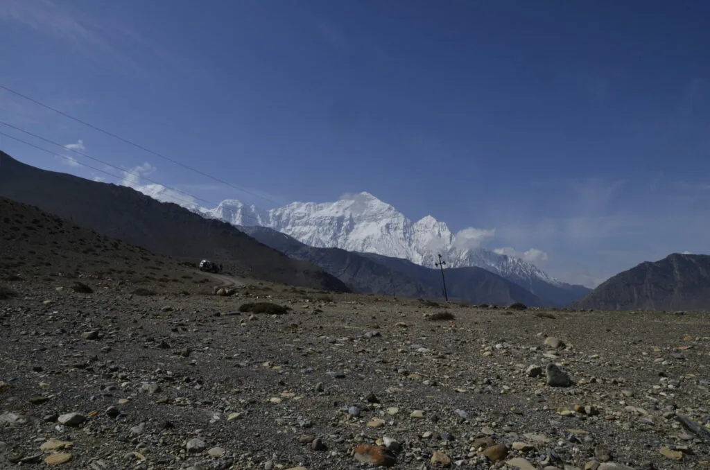 Snow-capped mountain peak visible across barren rocky terrain with power lines and a vehicle in the distance under clear blue sky
