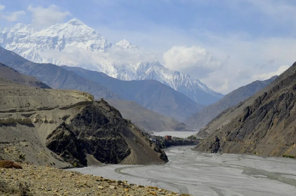 Snow-capped mountains frame a glacier-fed river flowing through a high-altitude valley with barren terrain and rocky outcrops