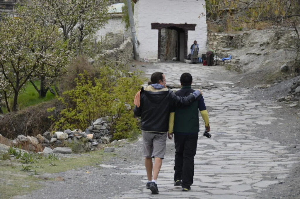 Two people walking away on a stone path toward a traditional building with dark wooden doorway in a mountainous village setting