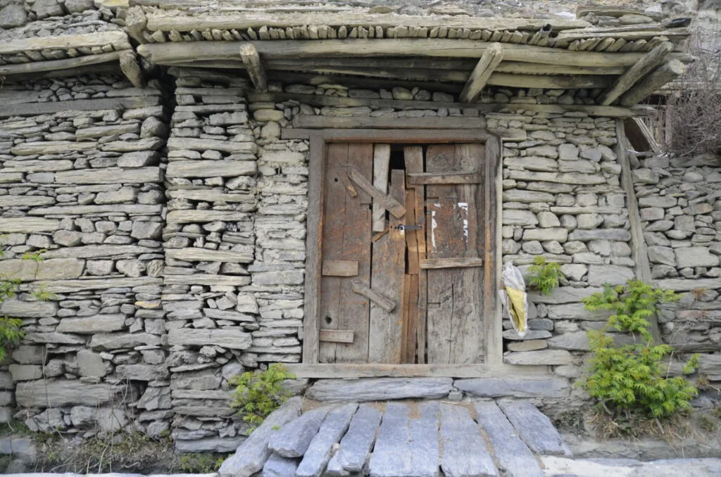 Weathered wooden door set in stacked slate stone wall of rural mountain building with wooden roof beams and moss-covered stone threshold