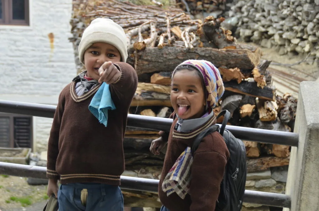 Two young children wearing brown sweaters and knit hats stand in front of stacked firewood, one pointing at camera with tongue out