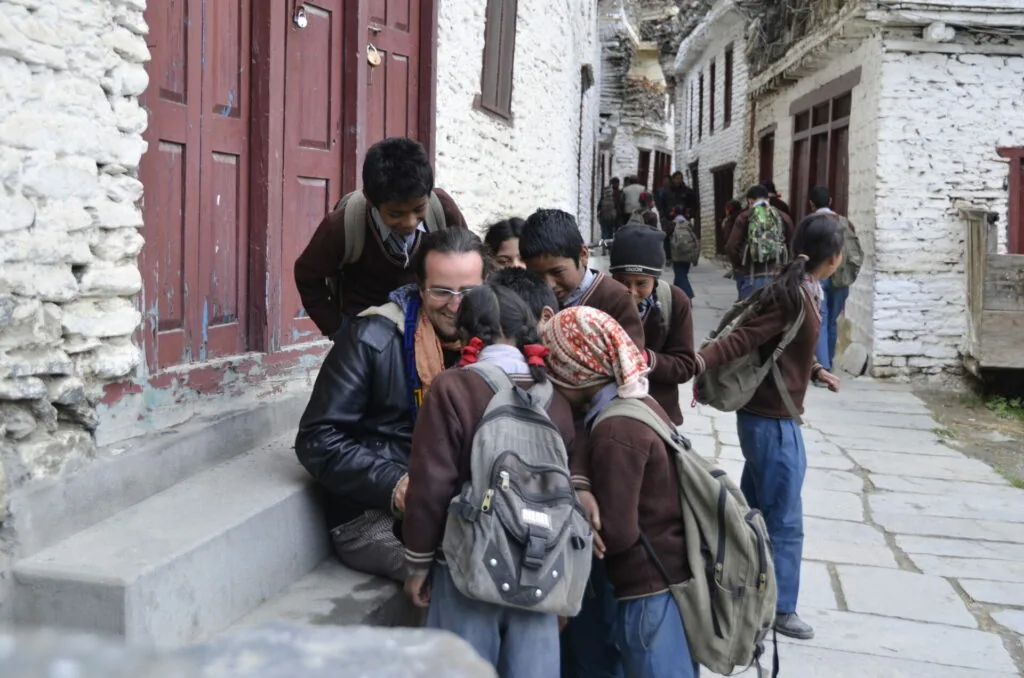 Adult woman crouching with group of children wearing backpacks on whitewashed stone street with red wooden doors