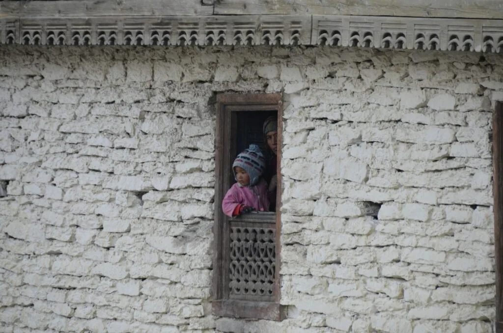 Person wearing blue hat and pink shirt visible in wooden-framed window of whitewashed brick building