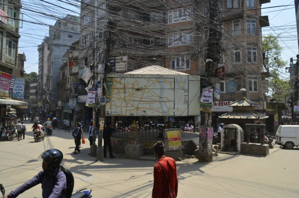 Busy urban street intersection with multi-story buildings, tangled power lines, a small shrine structure, and pedestrians walking near a white commercial structure with a faded map display