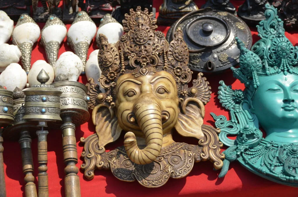 Golden Ganesha head sculpture displayed with ornamental bells, turquoise deity statue, and decorative brass items on red fabric