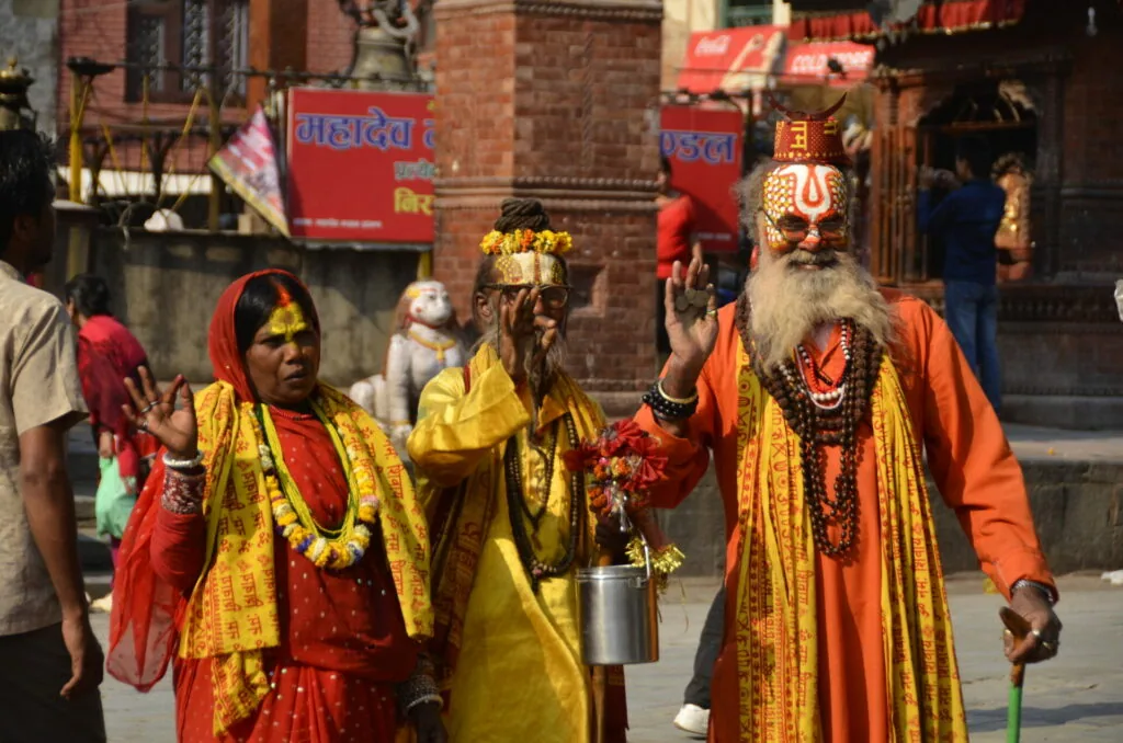 Three people in colorful traditional religious attire with face paint and garlands stand in a brick courtyard with red signage and other visitors in background.