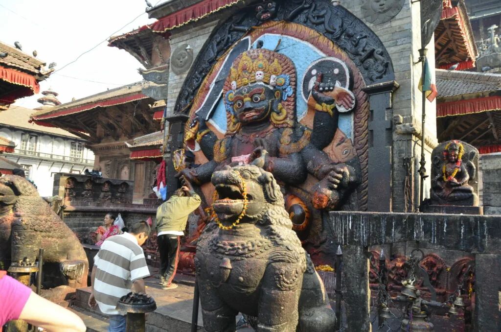 Colorful painted deity figure on temple wall with stone lion statue in foreground at Nepalese temple complex