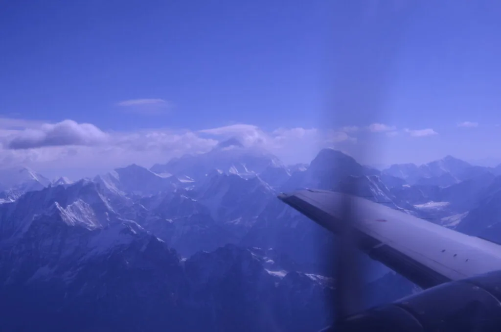 Airplane wing in flight over snow-capped mountain range under blue sky with clouds