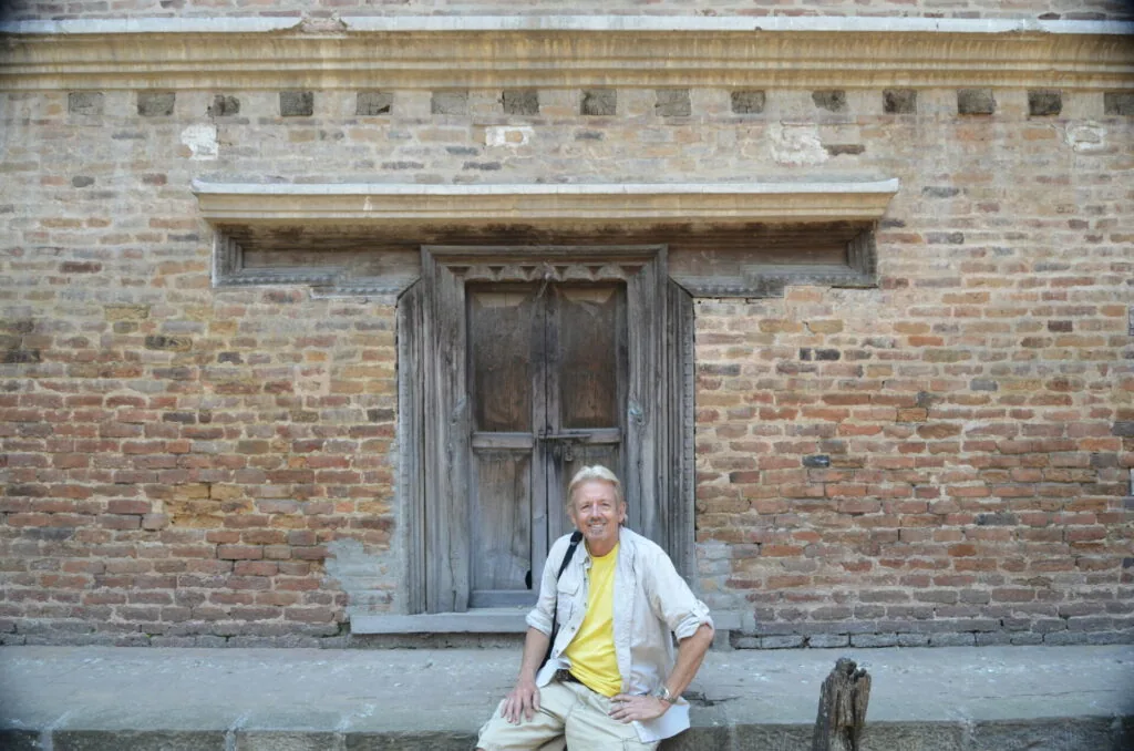 Man in yellow shirt and light jacket sitting in front of weathered brick building with wooden door
